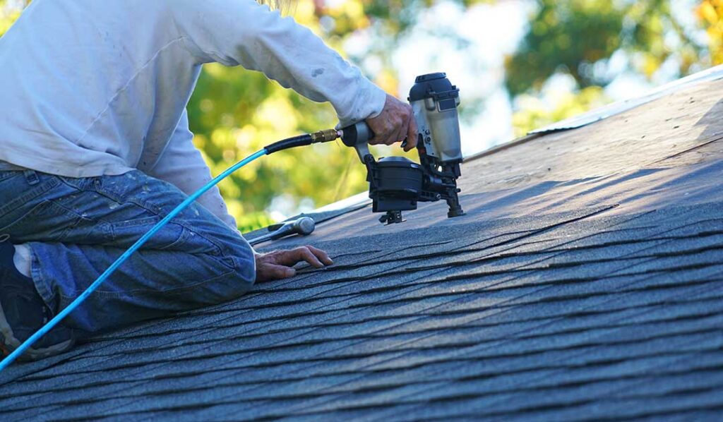 A homeowner in work boots and a harness steadies a ladder next to a house roof under clear daylight.