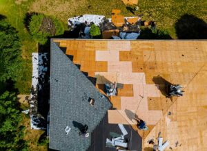 Roof replacement materials above a home while a worker inspects dry attic insulation and roof framing.
