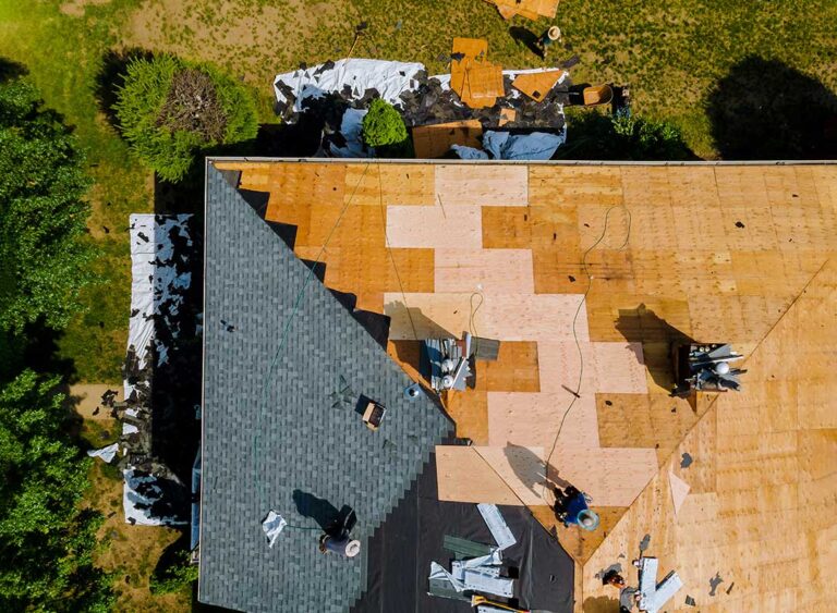 Roof replacement materials above a home while a worker inspects dry attic insulation and roof framing.